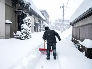 雪の積もった住宅街の路地で赤いスコップを使って雪かきをする男性